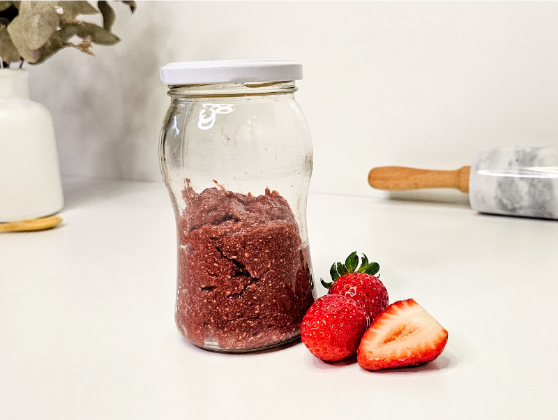 foto de un frasco de mermelada de frutilla sin azúcar baja en fodmaps sobre una mesada blanca, con un florero de fondo y una fresas al lado